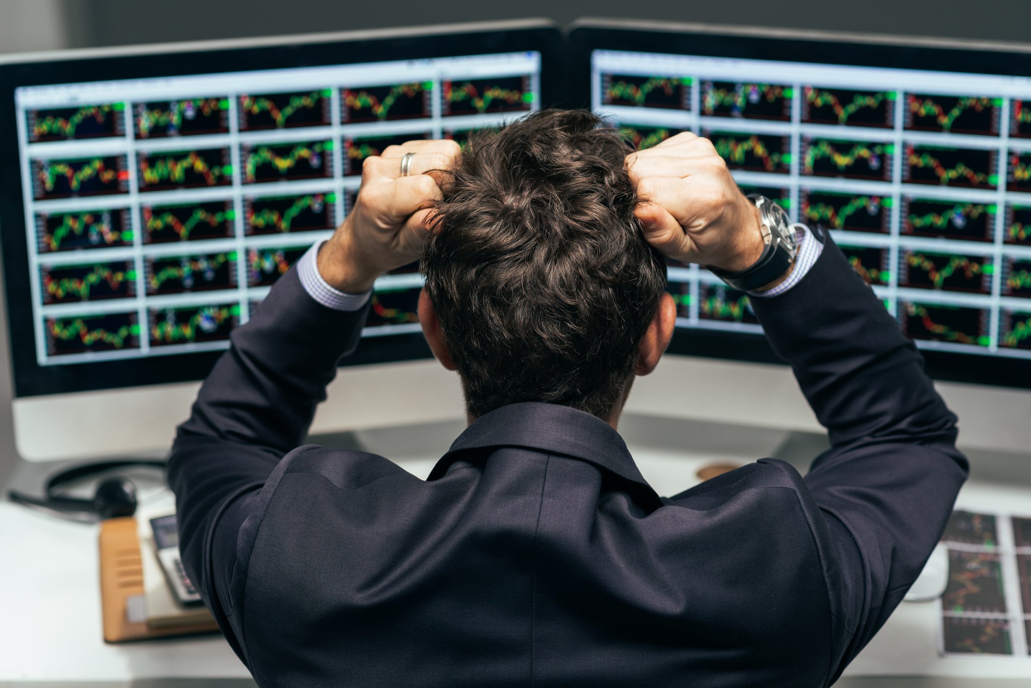 Man in front of monitors displaying stock charts, clenching fists to his head