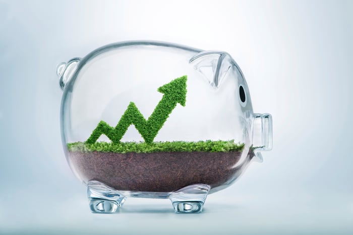 A glass piggy bank with an arrow made of grass growing out of a bed of soil.