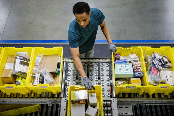 Amazon.com warehouse employee, seen from overhead, sorts yellow bins full of merchandise on a conveyor belt.
