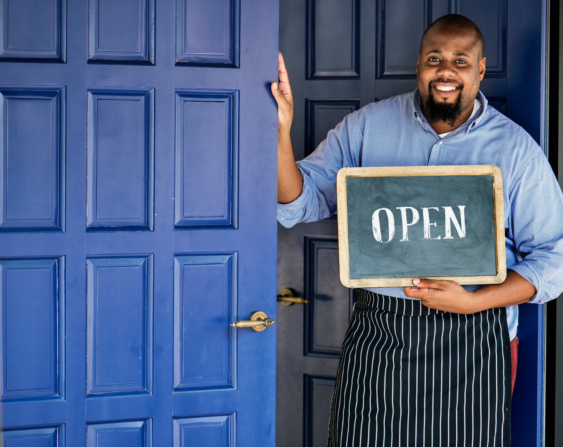 Man in apron standing next to blue door holding chalkboard with the word open written on it