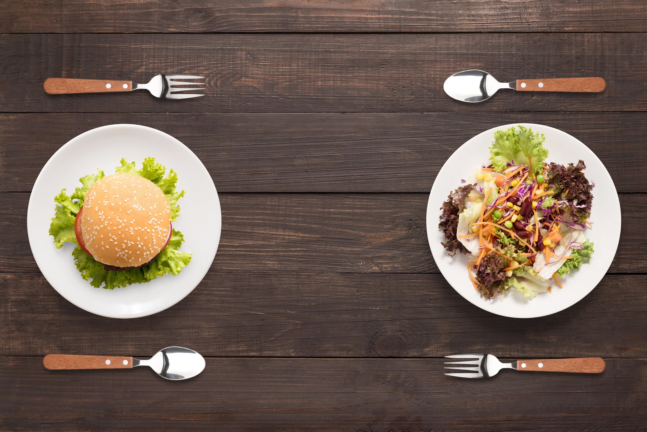 A burger on a plate flanked by a fork and spoon facing a salad on a plate flanked by a fork and spoon