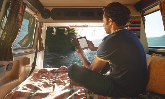 A young man sitting on the bed in the back of a van reading a Kindle.