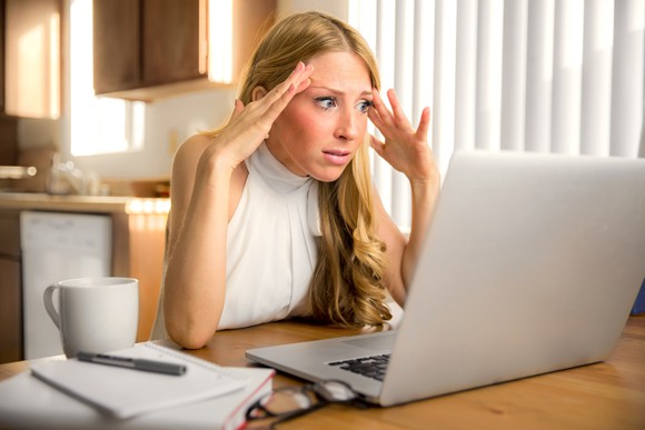 Woman at laptop holding her hands to her head and staring as if in shock