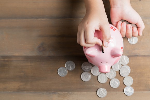 A child puts a coin in a pink piggy bank with other coins scattered around a tabletop.