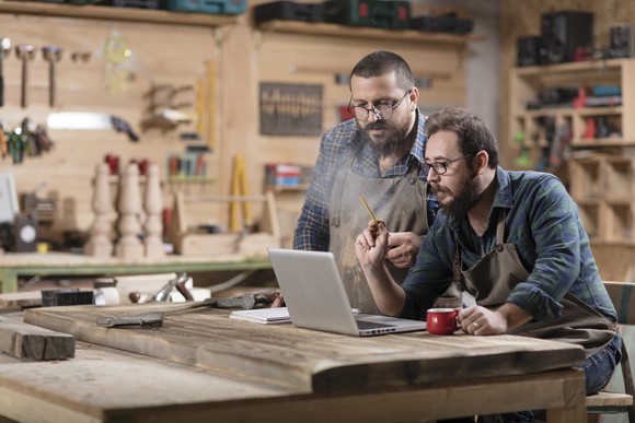 Two men in a workshop look at the screen of a laptop computer.