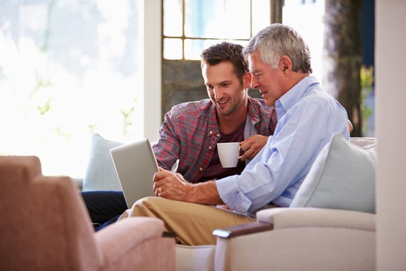 An older and younger man site side by side on a sofa, smiling as they look at the screen of a tablet.