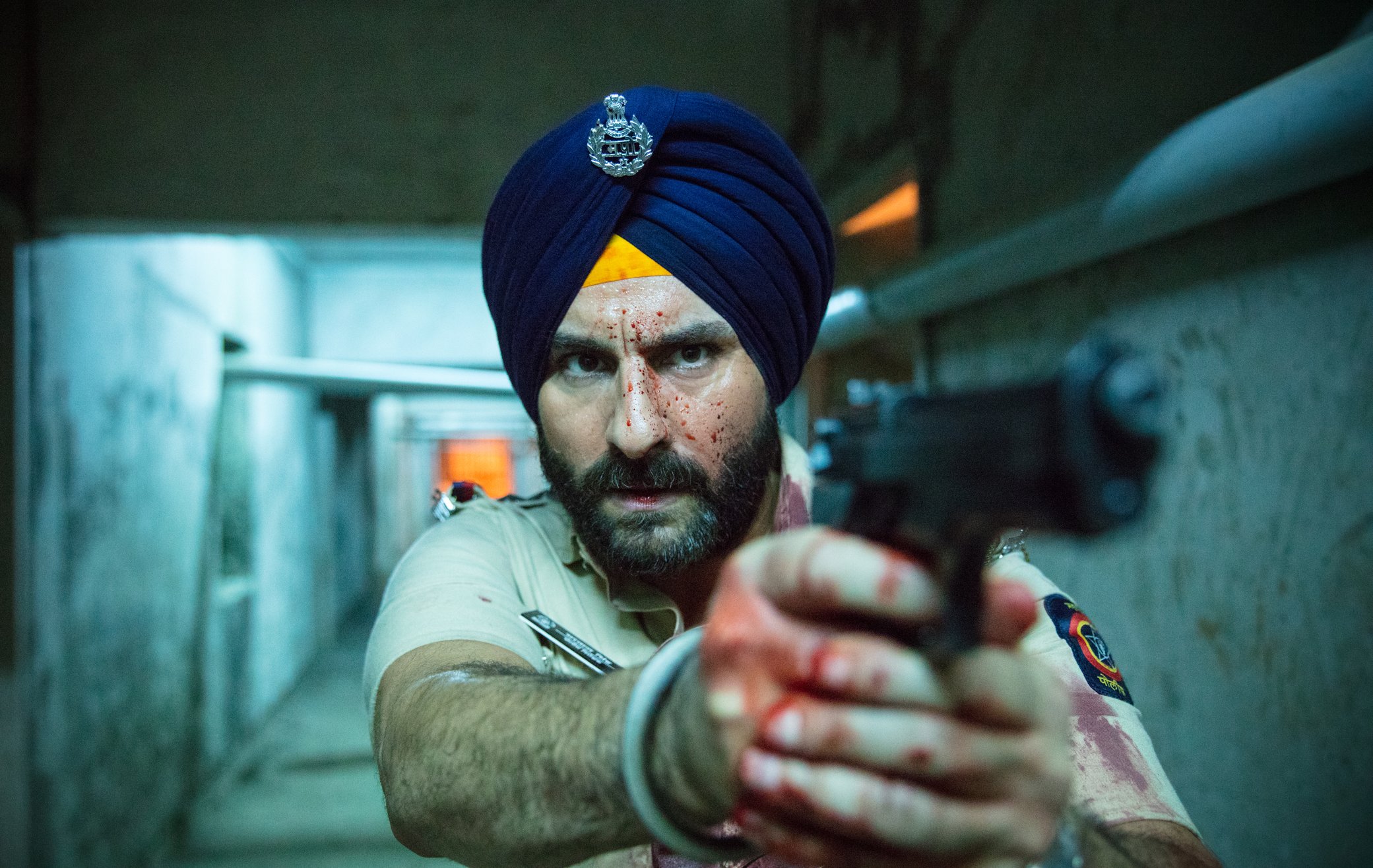 A man wearing a turban pointing a gun with his face and hands covered in blood spatter.