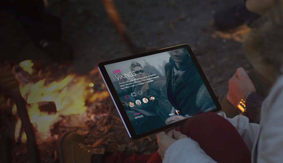 A person holding a tablet displaying a mockup of T-Mobile's TV app while sitting next to a campfire.