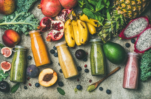 An assortment of colorful bottled smoothies surrounded by fruit and vegetables on a wood table.