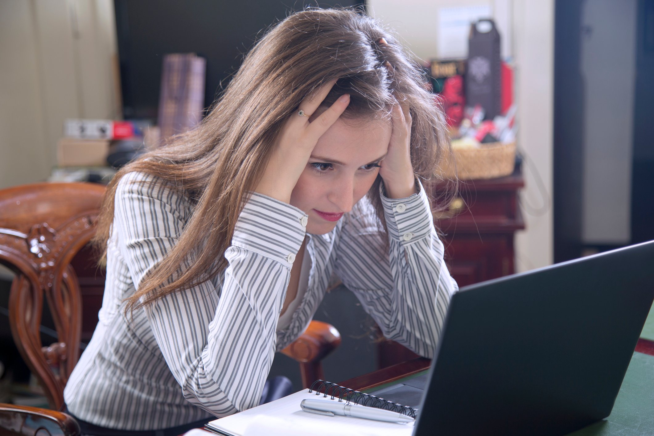 A frustrated woman grasping her head with both hands while reading news on her laptop.