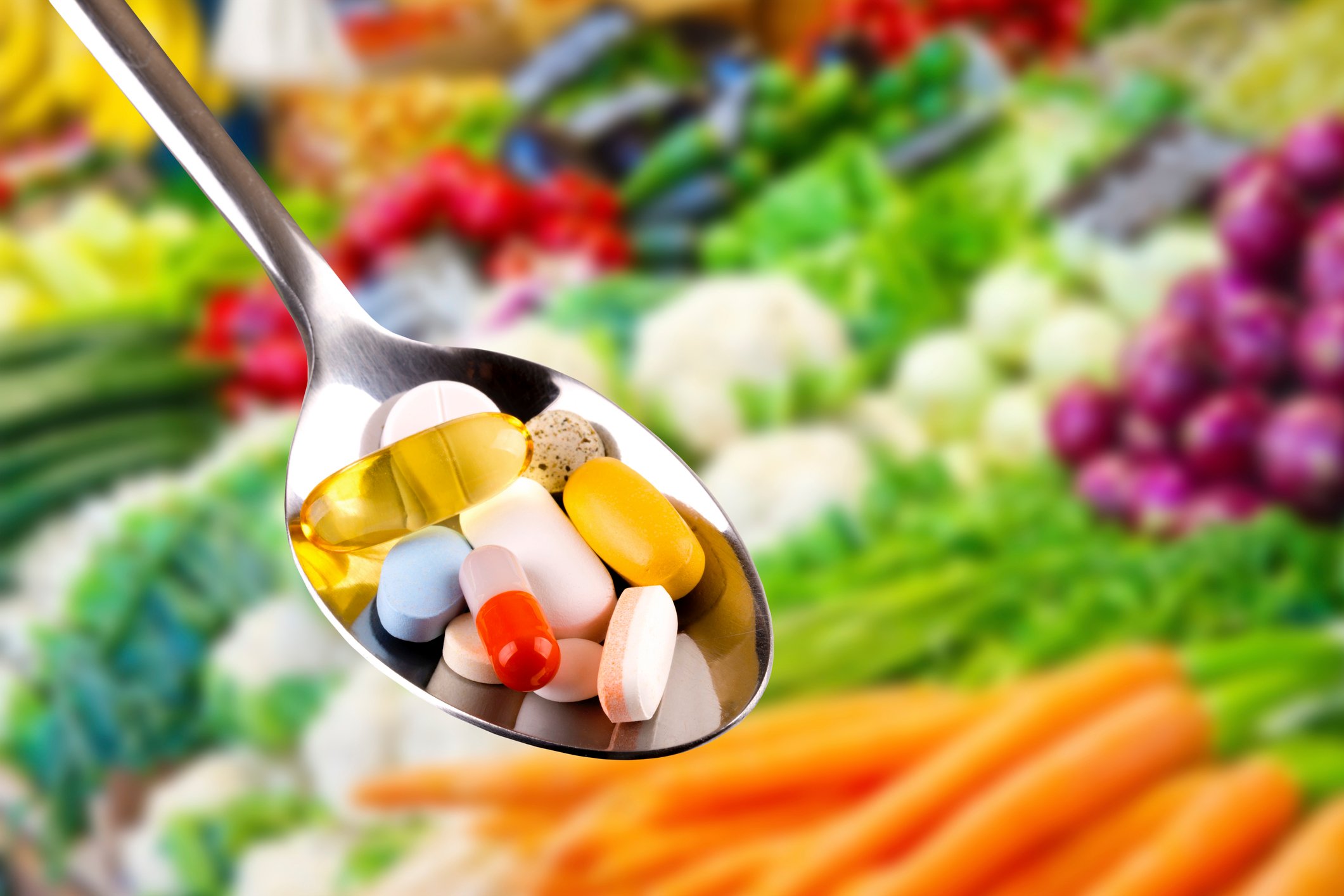 A tablespoon of pills with vegetables in the background.