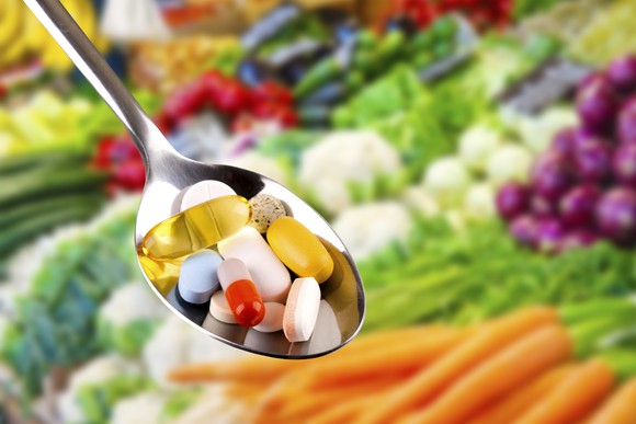 A tablespoon of pills with vegetables in the background.