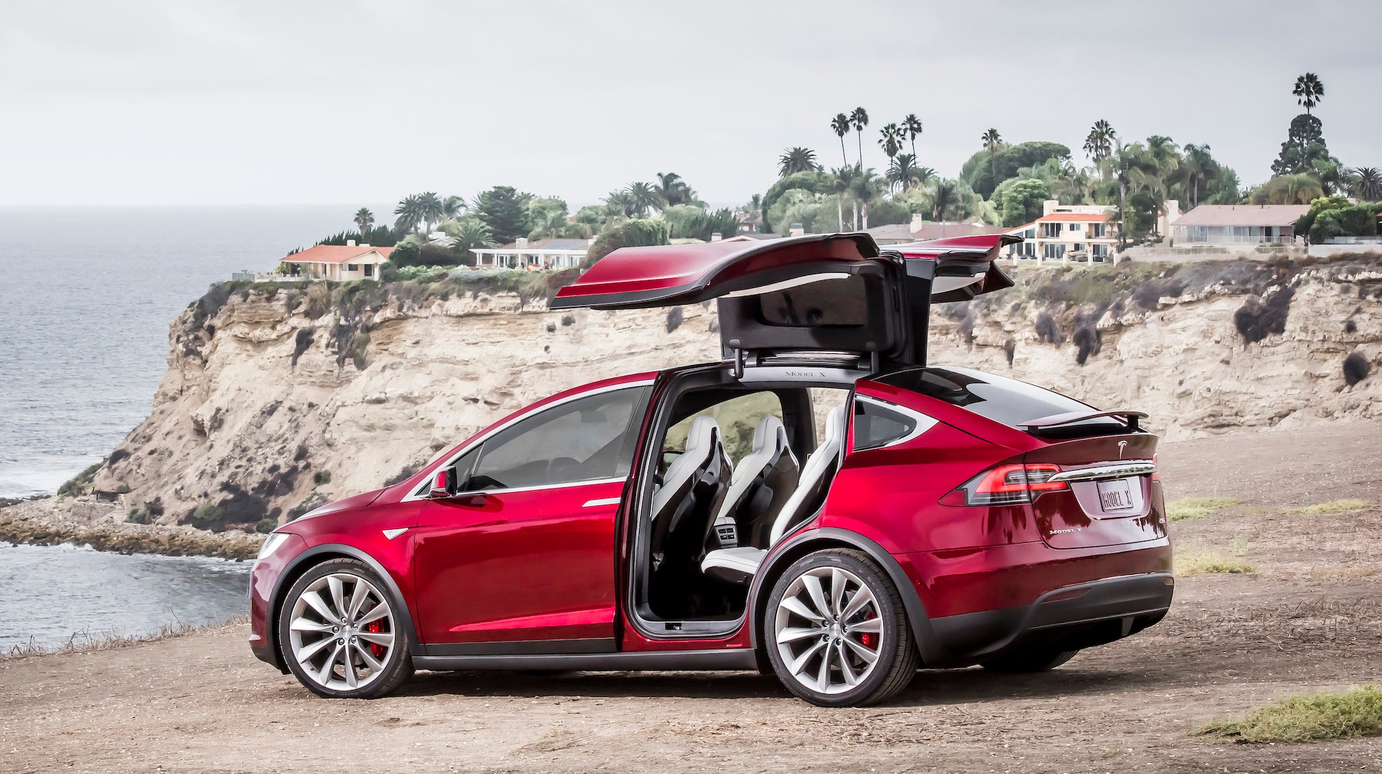 A red Tesla Model X SUV with its rear "falcon-wing" doors open, parked on a beach.