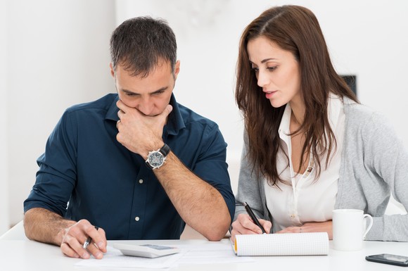 Man and woman at table; man has calculator in front of him while woman takes notes.