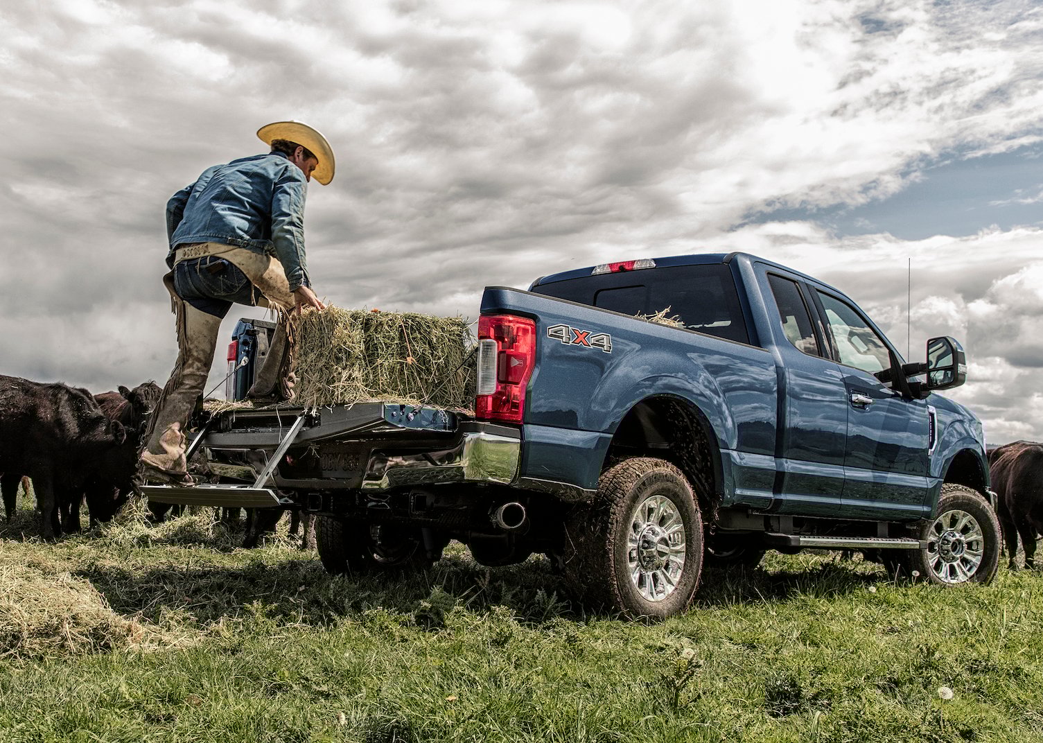 A rancher is shown unloading a hay bale from a blue 2019 Ford F-350 XLT, a heavy-duty full-size pickup. Cattle are visible in the background.