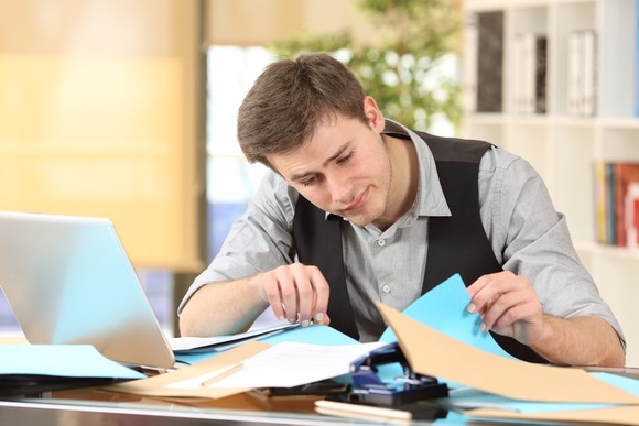 Man searching for something underneath folders on a messy desk.