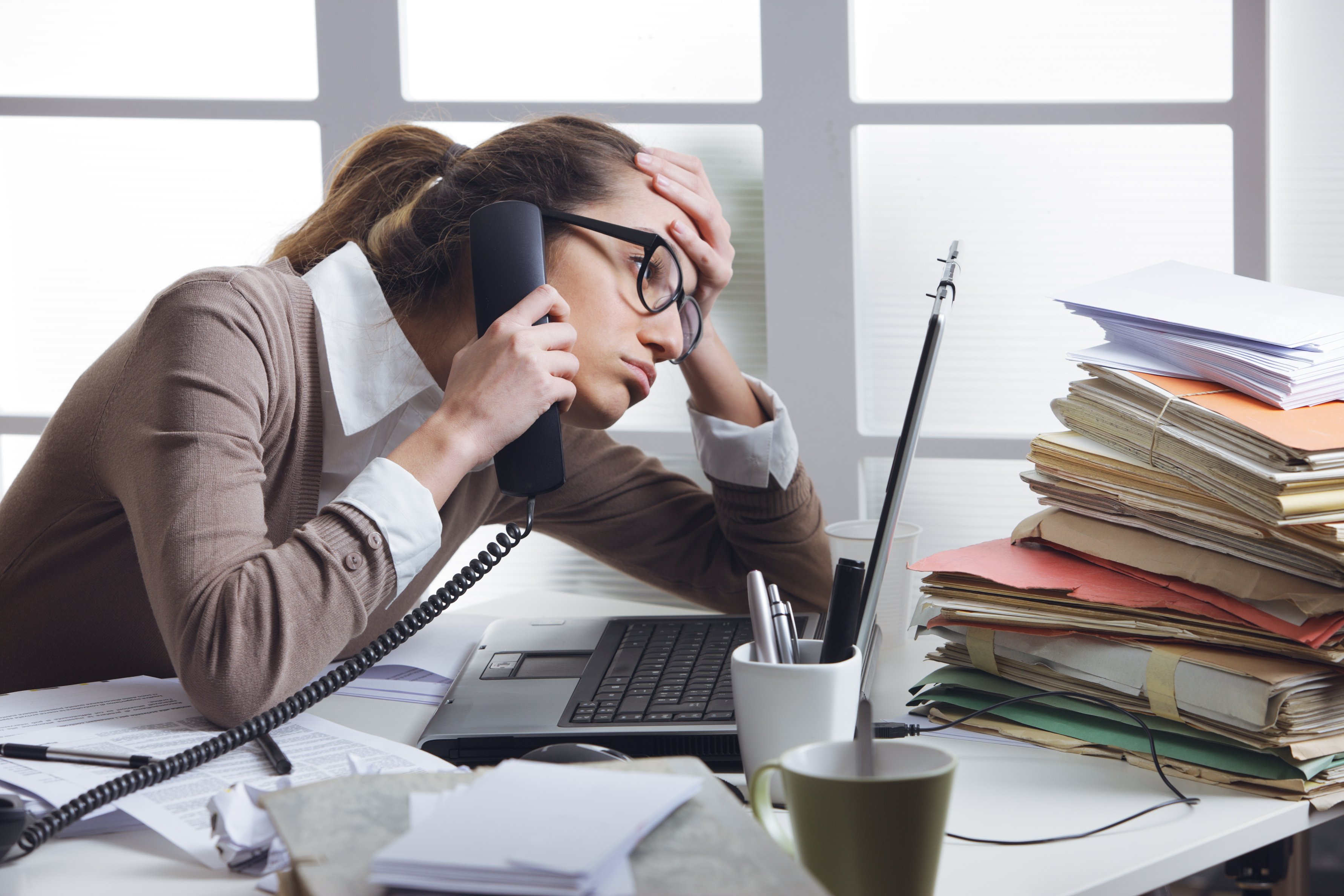 Professionally dressed woman on phone in an office, holding her head with bored expression.