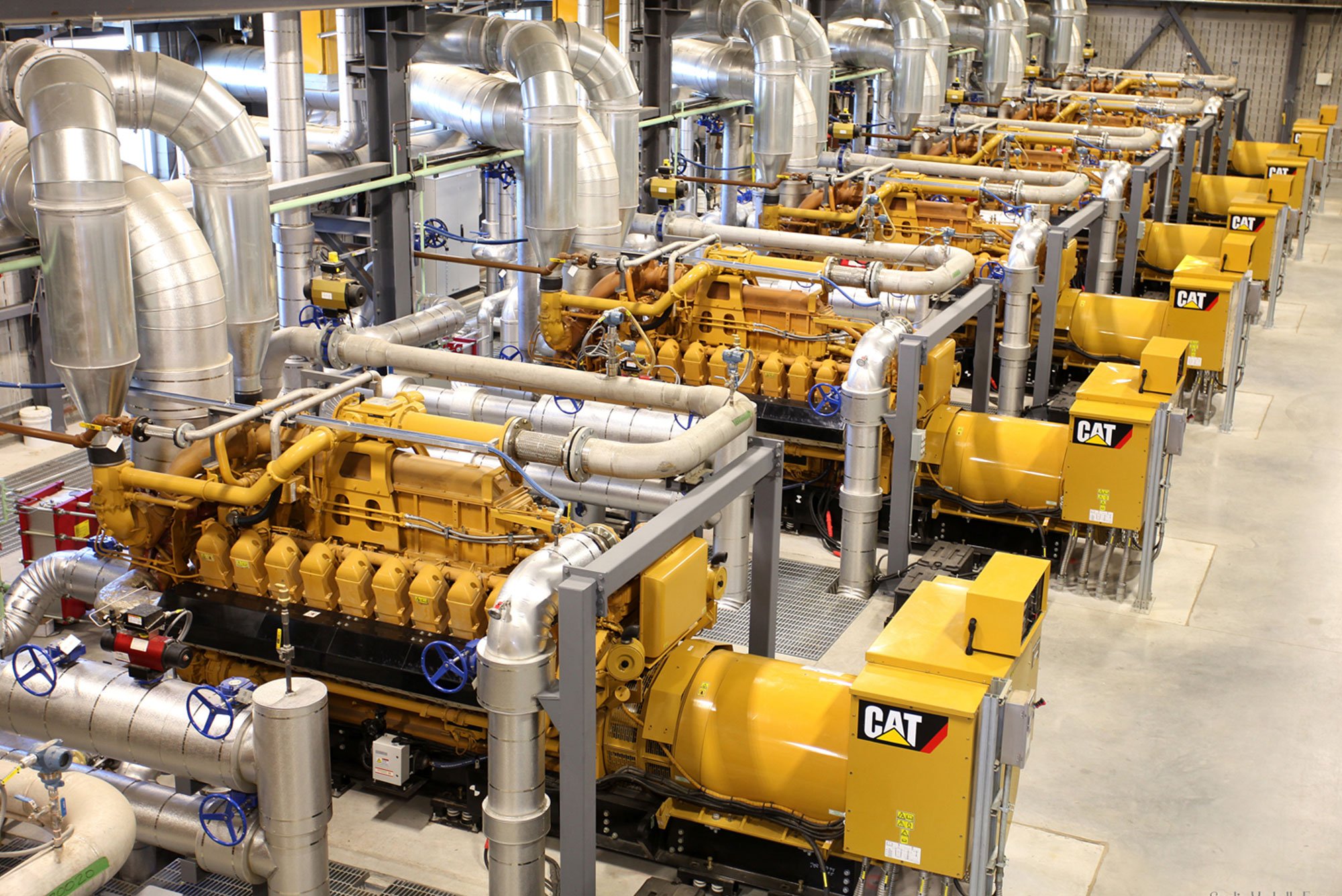 A row of Caterpillar engines in a factory environment attached to piping.