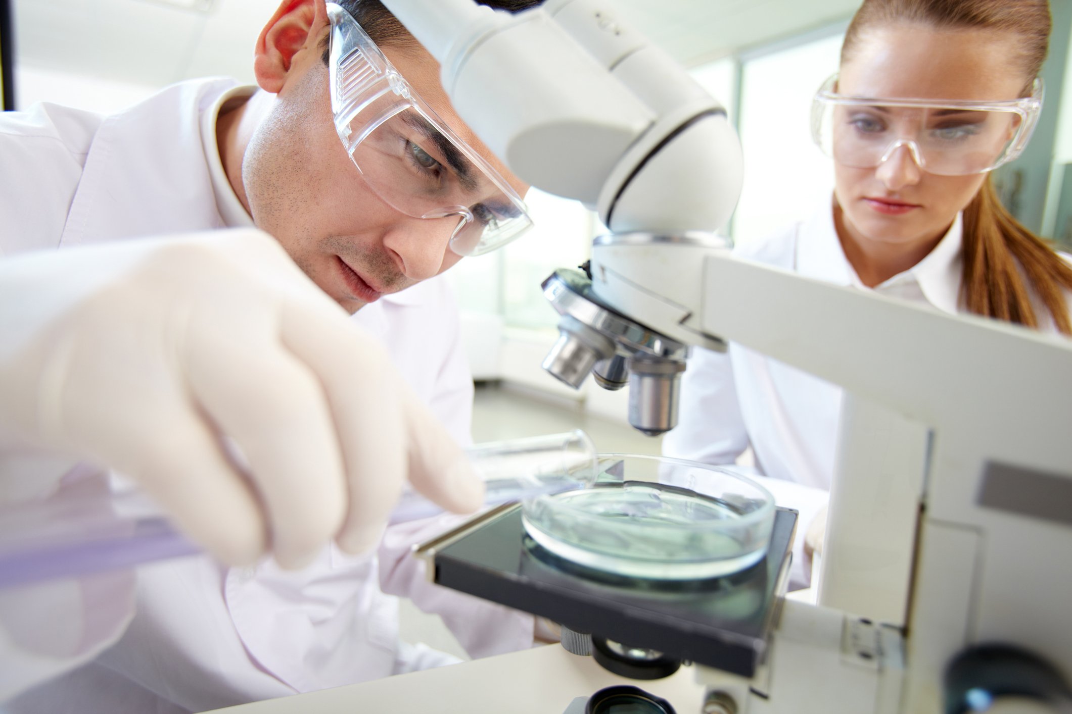 A man and a woman wearing lab coats and protective glasses looking at a petri dish under a microscope