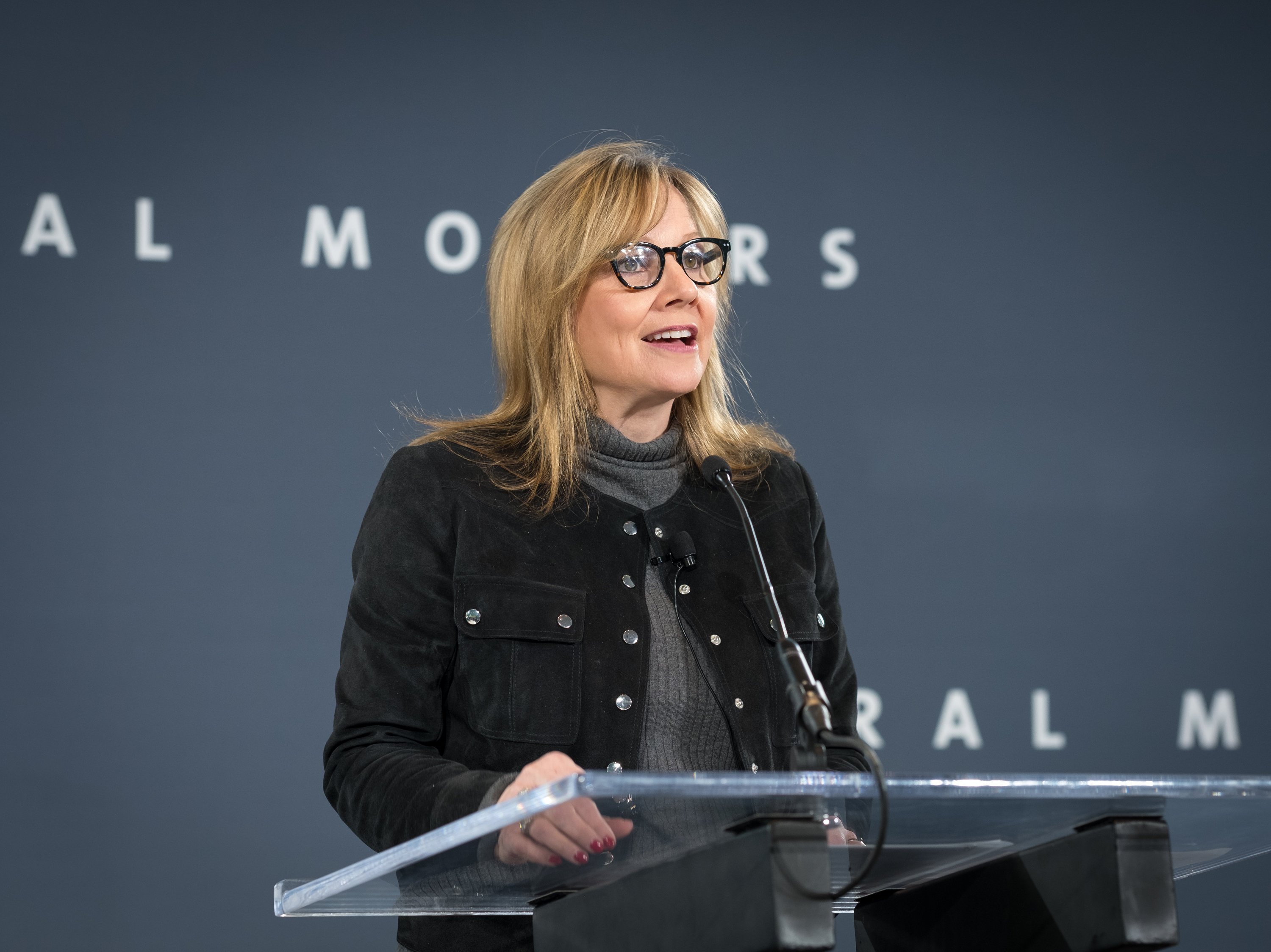 GM CEO Mary Barra standing at a podium in front of a dark-blue General Motors backdrop.