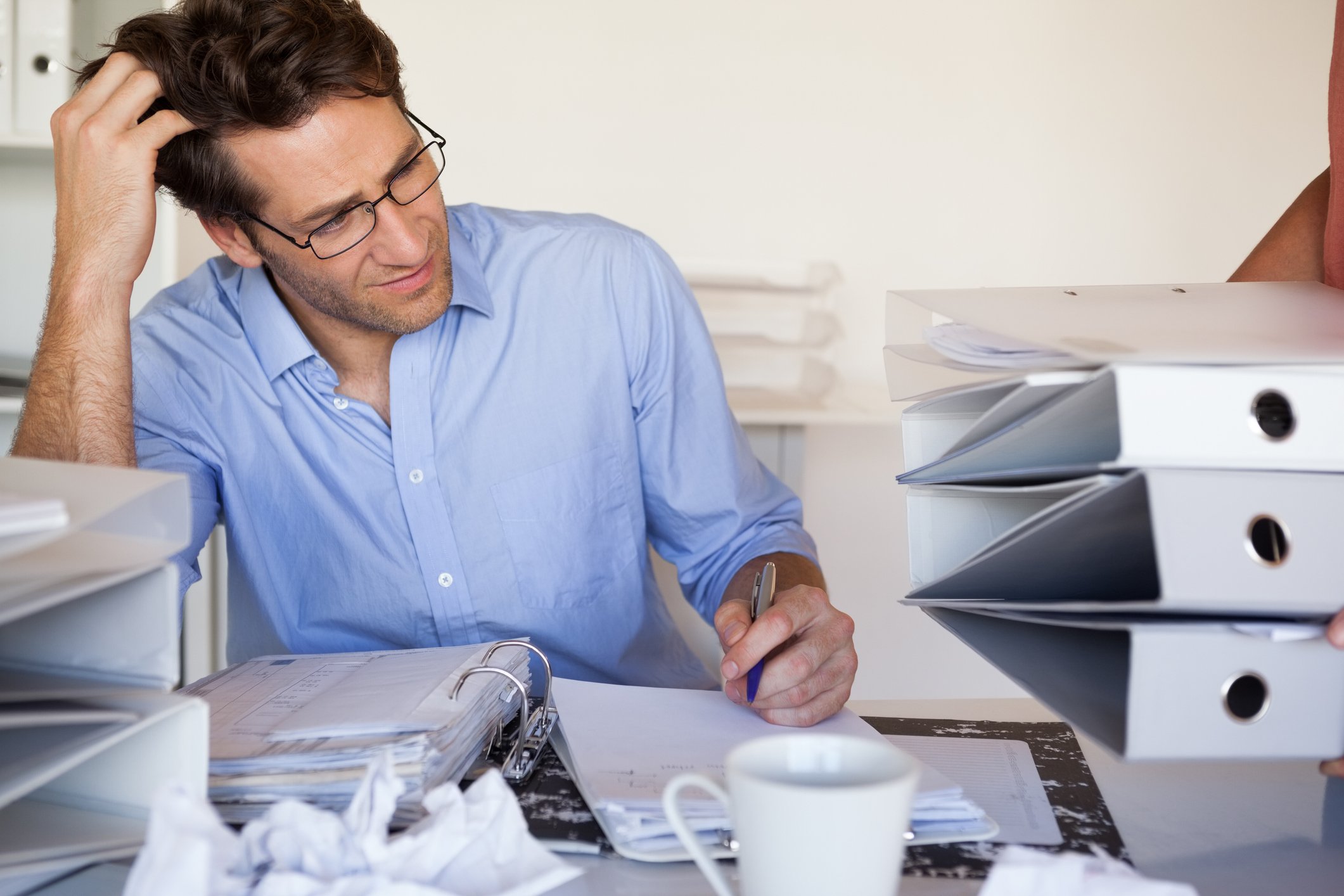 Man at cluttered desk scratching his head while taking notes