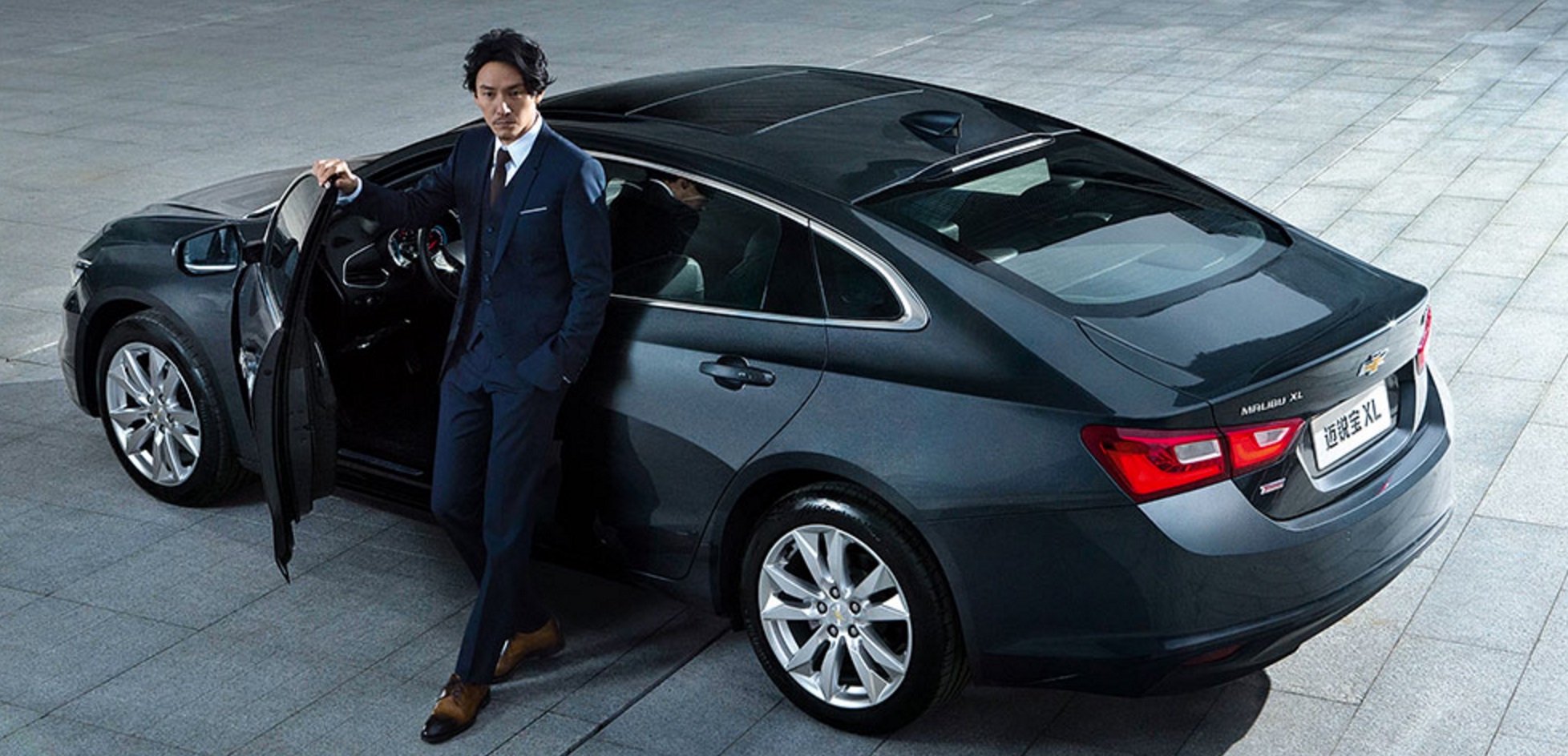 A man in a suit stands next to a dark gray Chevrolet Malibu XL, a large midsize sedan.