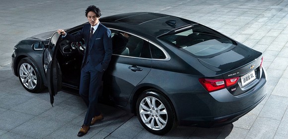 A man in a suit stands next to a dark gray Chevrolet Malibu XL, a large midsize sedan.