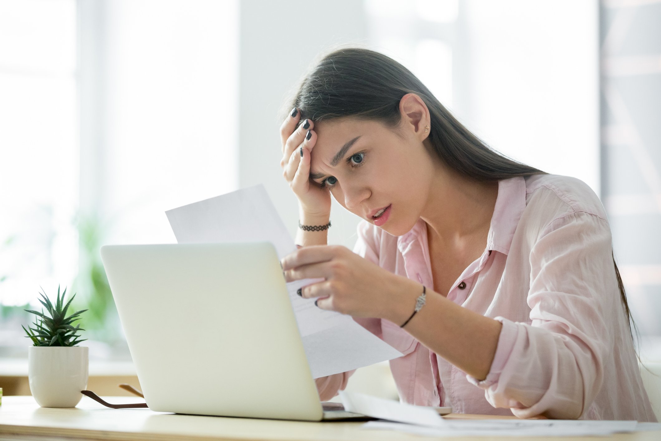 Woman at laptop looking at sheet of paper with stressed expression.