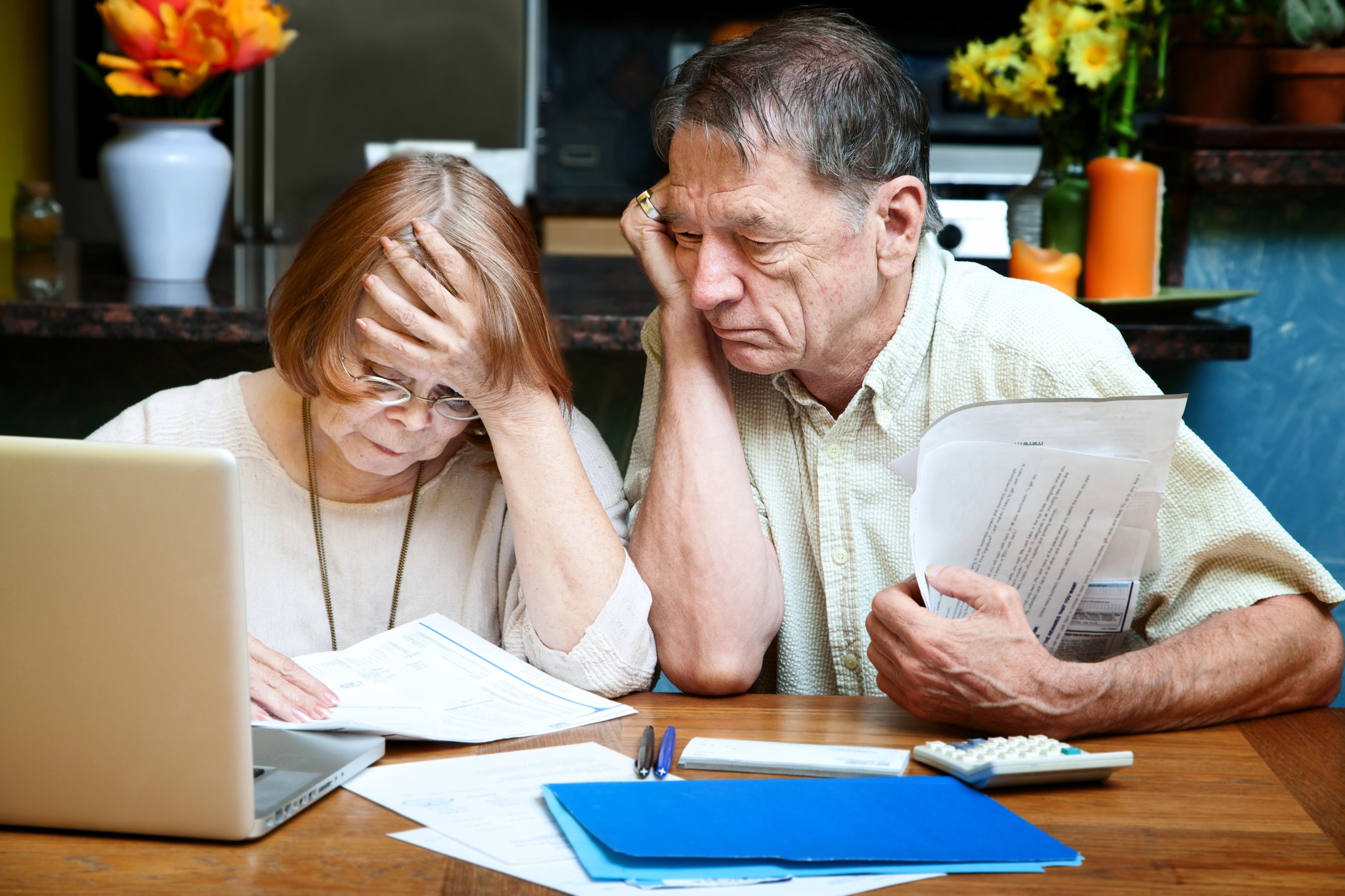 A senior couple, gathered at a desk in front of a computer, look concerned as they examine piles of papers.