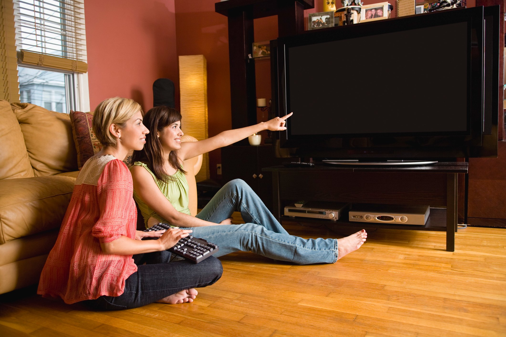 Two women sit on the floor of a living room, while one points to a large TV screen. 