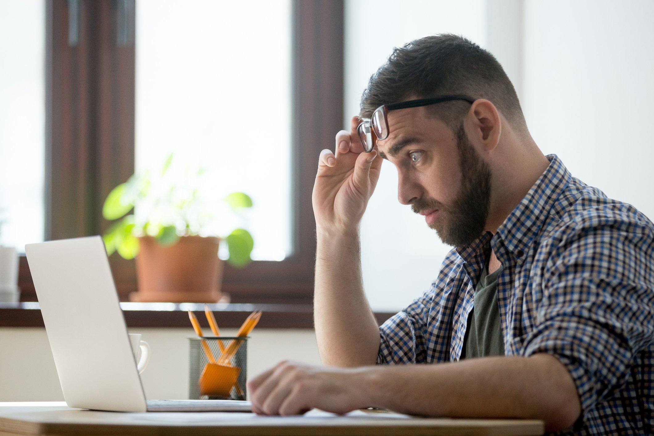 Worried man raising eyeglasses from his eyes while looking at his laptop.