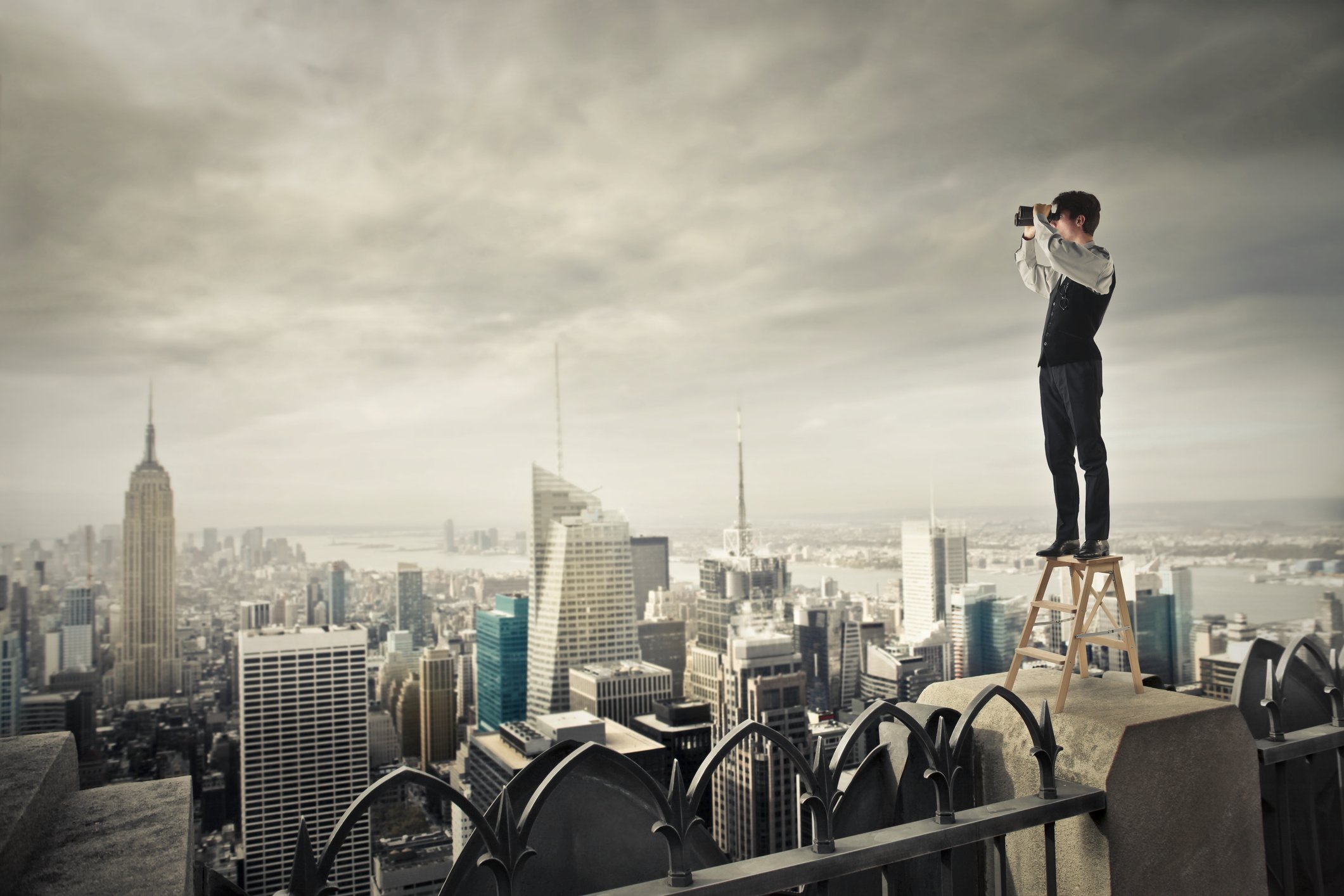 Man standing on step stool on top of skyscraper looking through binoculars