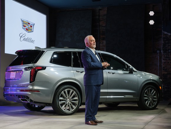 Carlisle is speaking on a stage. Behind him is a silver Cadillac XT6, a midsize luxury crossover SUV.