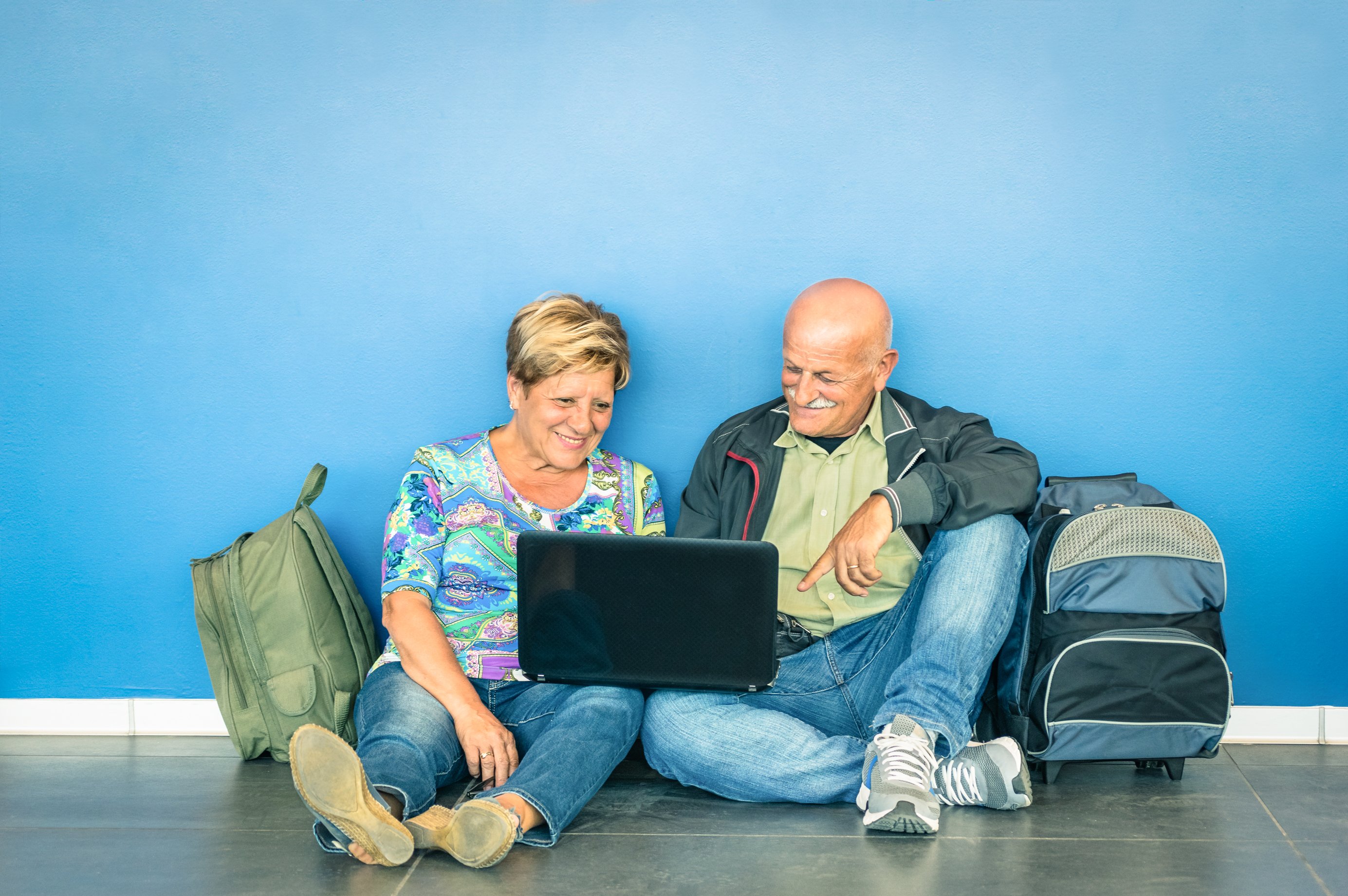 A senior couple sitting on the floor, looking at a laptop, with backpacks beside them.