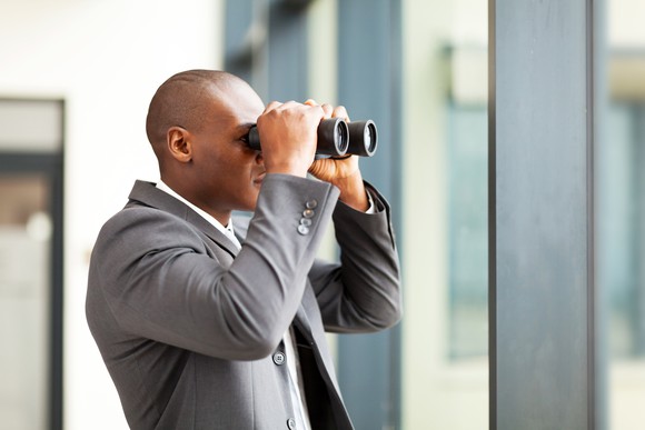 A bald businessman looking out a window with binoculars.