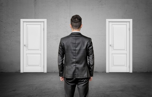 A businessman with his back turned stands between two identical closed white doors.
