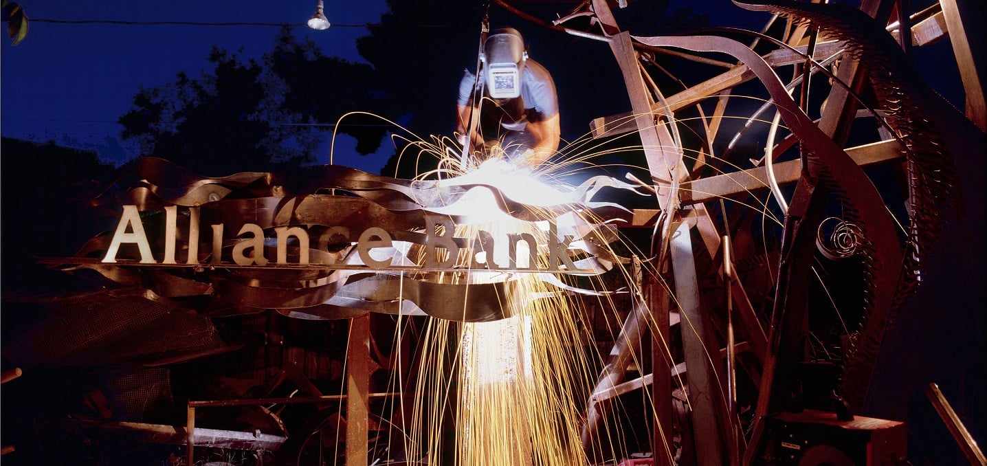 Welder working on metal sculpture with Alliance Bank name incorporated into it.