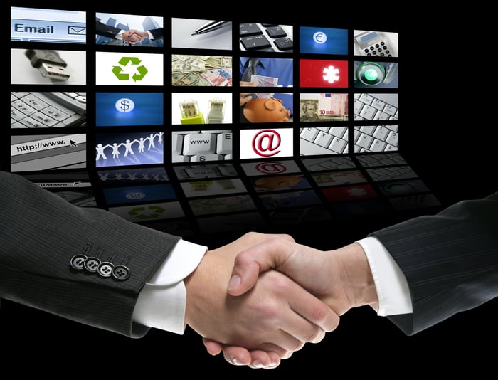 Two businessmen shake hands in front of 24 large TV screens showing various types of content.