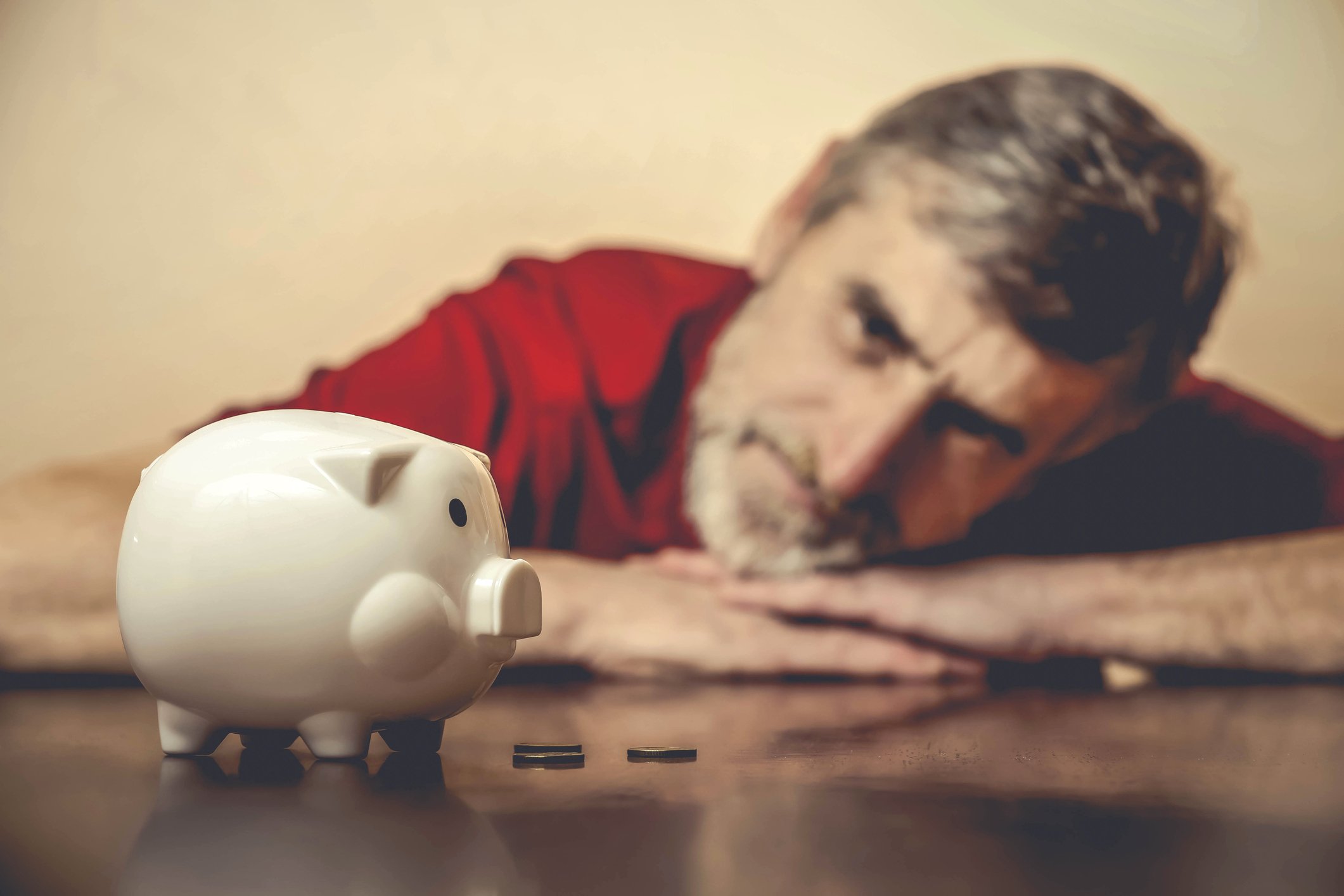 A worried baby boomer lying his head and arms on a table while looking at a mostly empty piggy bank.