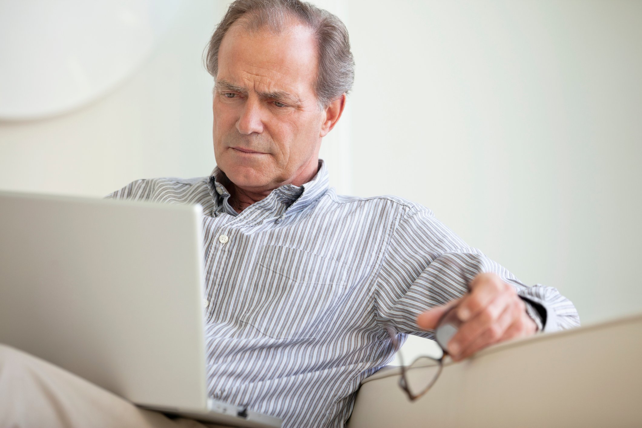 A baby boomer sitting on the couch and holding his glasses while critically looking at information on a laptop. 