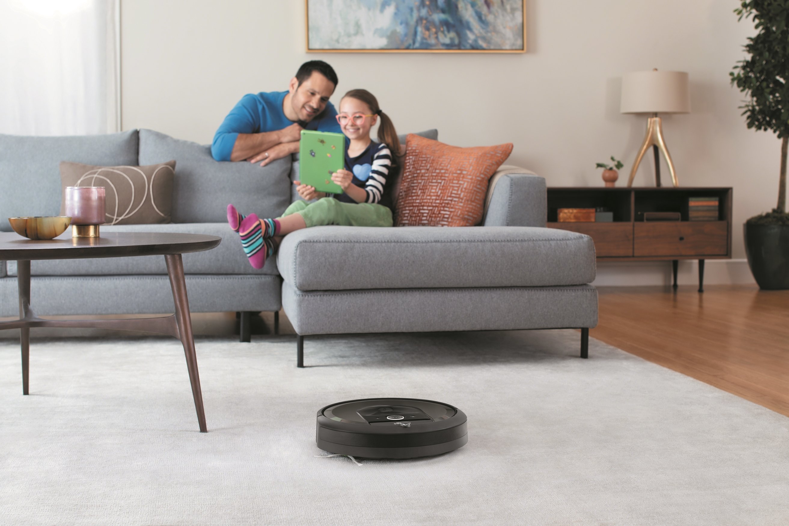A young girl on a couch, while her father leans over the back, while a robotic vacuum cleans a carpet in the foreground.