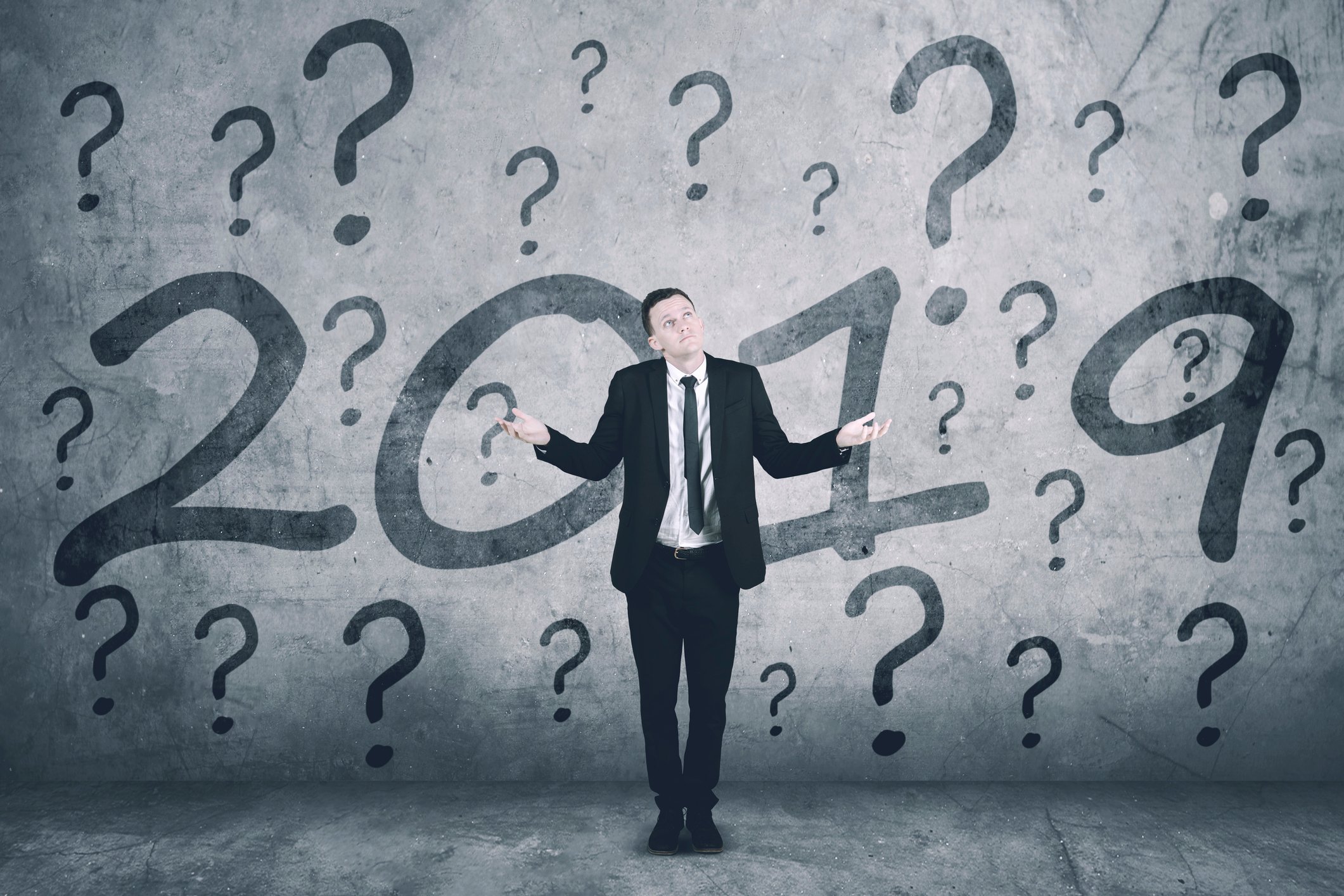 Man in a suit holding out hands while standing in front of a wall with 2019 and question marks painted on it