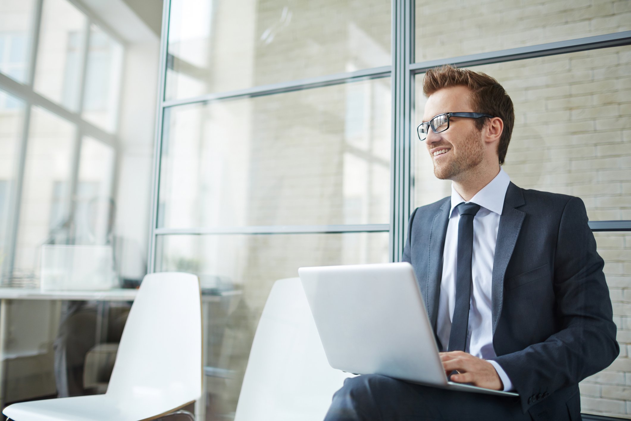 Smiling young businessman sitting in a chair and using laptop in a lobby.