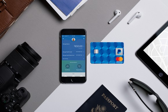 A smartphone and a PayPal branded credit card on a desk surrounded by a camera, passport, sunglasses and earbuds.