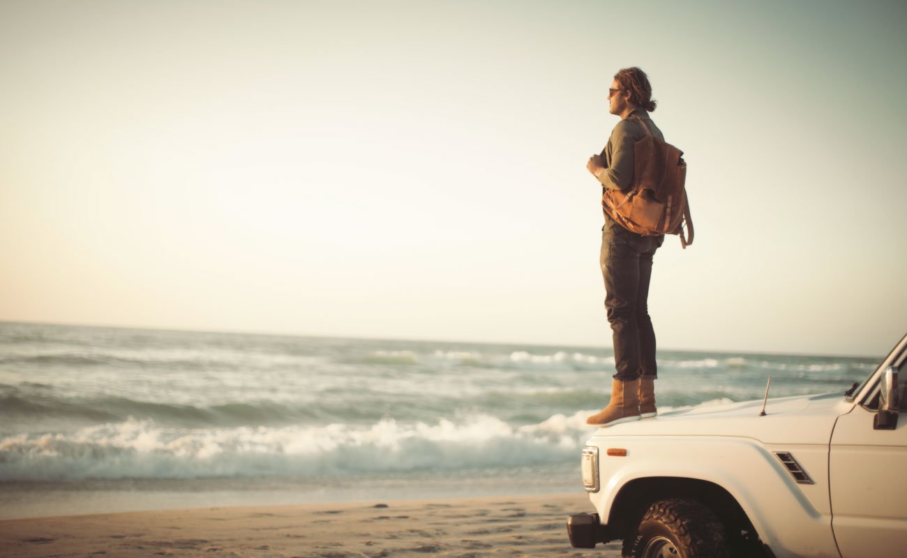 A man in Ugg boots stands on the hood of a truck on the beach.