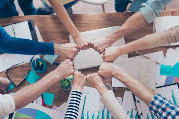 A group of workers fist bumping over a table covered with papers and business items.