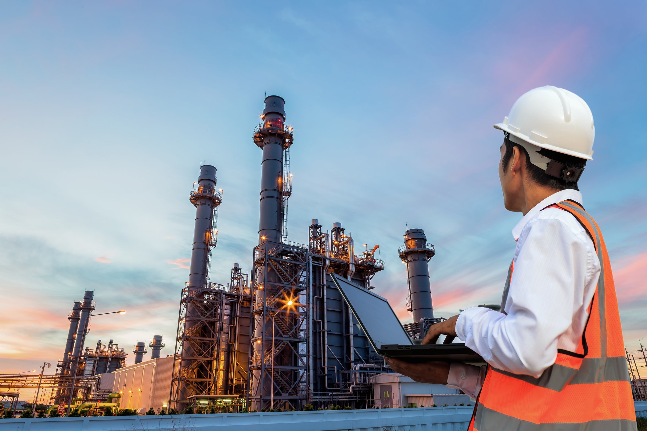 Engineer with laptop stands before an oil refinery at dusk.