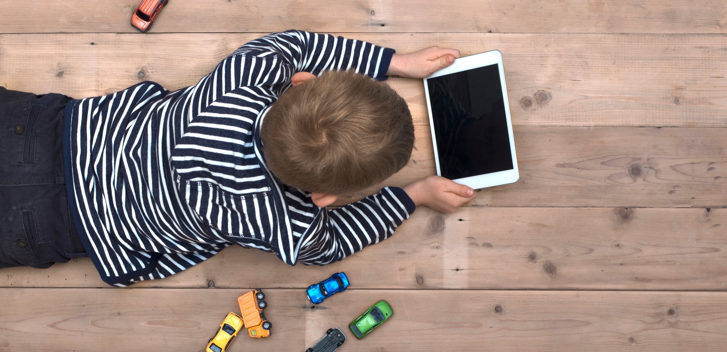 A young boy looks at a tablet computer while lying on the floor with toy cars scattered around him.