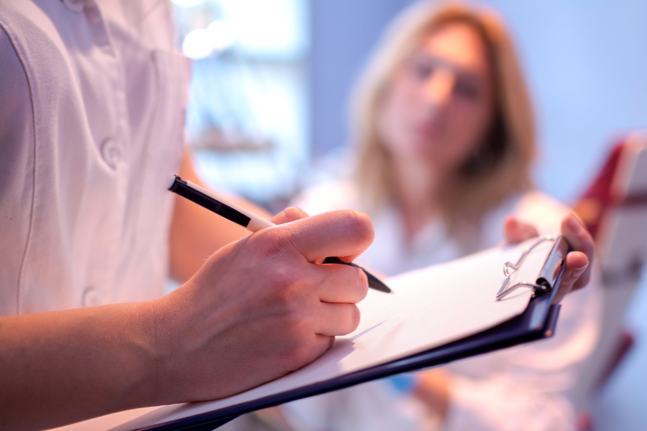 Doctor taking notes on a clipboard with a nurse in the background.