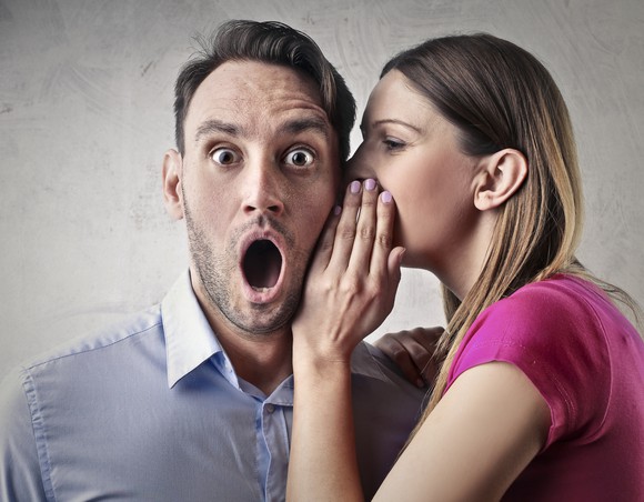 Woman whispering to a man whose mouth is wide open in surprise. 